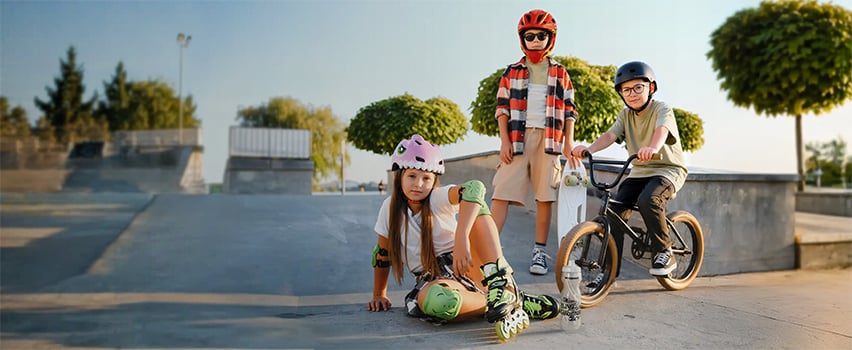Drei Kinder mit Helmen und Schutzausrüstung von Wozinsky auf Skateboard, Rollschuhen und BMX-Fahrrad im Skatepark